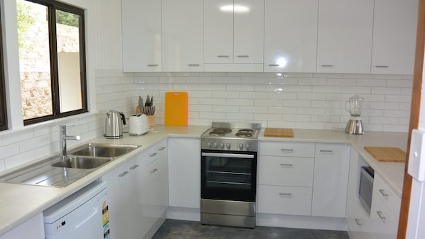 Een compacte keuken met witte kastjes, een roestvrijstalen kookplaat, een vaatwasser en een raam met uitzicht op een bakstenen muur.