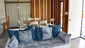 Een woonkamer met een witte bank, blauwe kussens met patroon, een houten eettafel met witte stoelen en een plafondventilator.