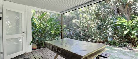 A covered patio with a table and bench, surrounded by lush greenery.