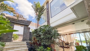 A modern hotel interior with a staircase leading to a balcony, surrounded by lush greenery and a view of the ocean. - Villa Amanie - Modern Balinese Luxury with Spectacular Views (Balangan)