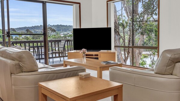 A modern living room with a large TV, comfortable seating, and a scenic view through large windows. - Coolabah Court - Merimbula (Merimbula)