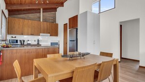 A modern kitchen with wooden cabinets, a central island, and a dining area with a wooden table and chairs. - Coolabah Court - Merimbula (Merimbula)