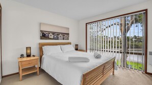A bedroom with a wooden bed, a nightstand with a lamp, a painting above the bed, and a view of the outdoors through a sliding door. - Coolabah Court - Merimbula (Merimbula)