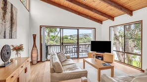 A spacious living room with a large sofa, a coffee table, and a TV on a wooden stand. There is a balcony with a view of trees and hills. - Coolabah Court - Merimbula (Merimbula)