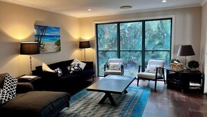 A living room with a dark-colored sofa, two armchairs, a coffee table, and a large window with a view of tropical foliage. - 7 Belle Court - Rainbow Shores. Huge Beach House! Pets Welcome. Property not fenced. Ducted aircon (RAINBOW BEACH)