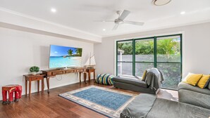 A living room with a TV, wooden furniture, a sailboat model, a large window with a view of greenery, and a ceiling fan. - 7 Belle Court - Rainbow Shores. Huge Beach House! Pets Welcome. Property not fenced. Ducted aircon (RAINBOW BEACH)
