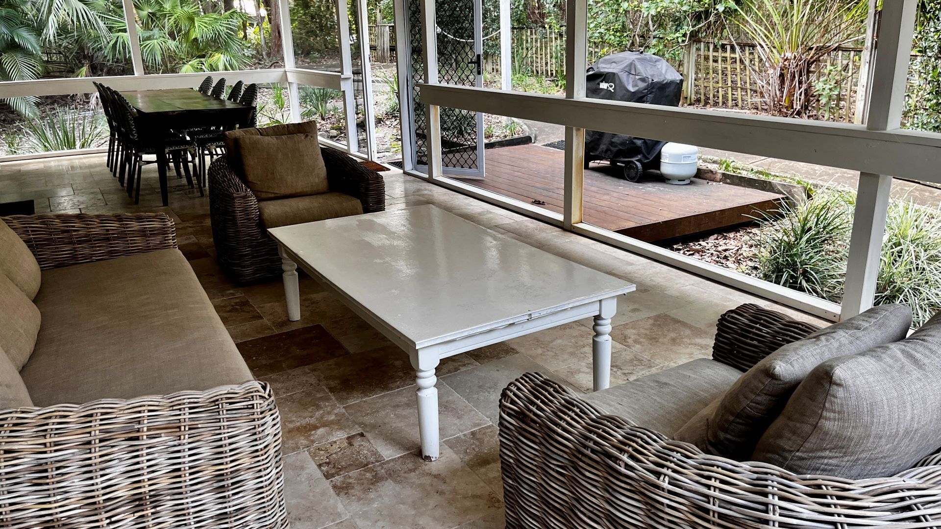 A sunlit patio area with wicker furniture, a white table, and a view of a garden and barbecue grill.