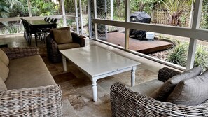 Een zonnig terras met rieten meubilair, een witte tafel en uitzicht op een tuin en een barbecue.