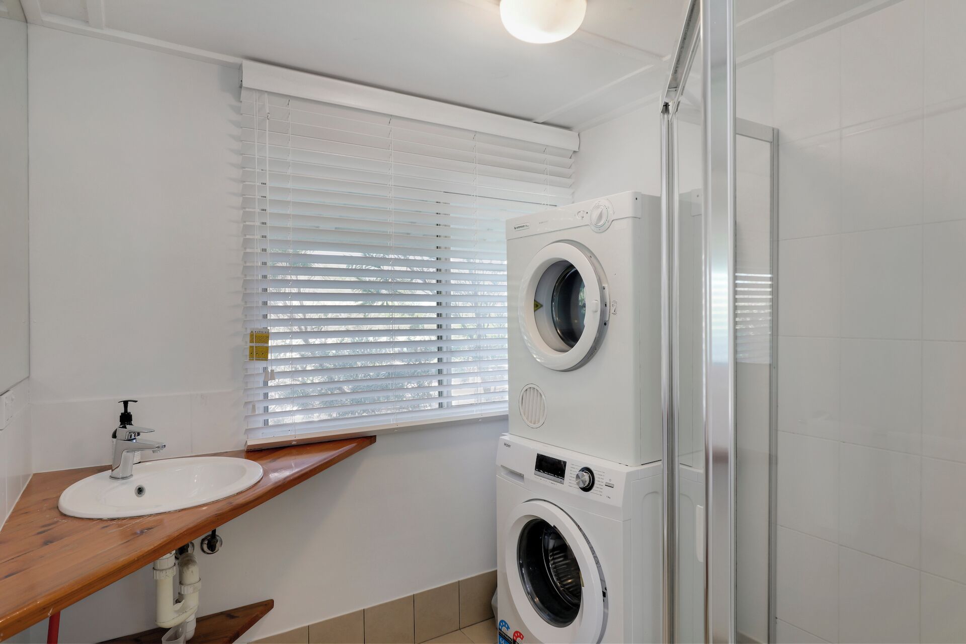 A compact bathroom with a sink, two stacked washing machines, and a shower area.