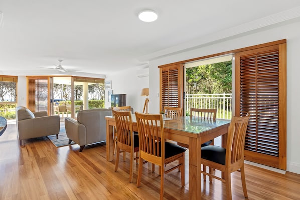 A dining area with a glass table and wooden chairs, large windows with blinds, and a ceiling fan.