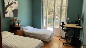 Une chambre avec deux lits, un bureau avec un ordinateur et une fenêtre donnant sur des arbres.