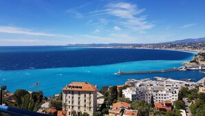 Un paysage urbain côtier avec un bâtiment imposant doté d’un toit recouvert de tuiles rouges et de nombreuses fenêtres, situé près d’une promenade en bord de mer, avec une jetée et un phare.
