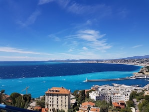 A coastal cityscape with a prominent building featuring a red-tiled roof and multiple windows, situated near a waterfront with a pier and a lighthouse. - NICE MOST AMAZING SEA + MOUNTAIN VIEWS ,2 A/C BEDROOMS,GARAGE FOR EV,3TERRACES (Nice)