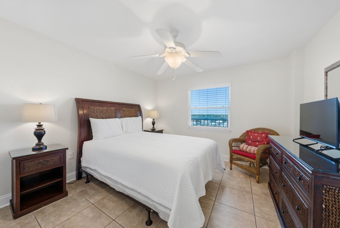 A bedroom with a bed, a ceiling fan, a window with blinds, a wicker chair, a dresser, and a television.