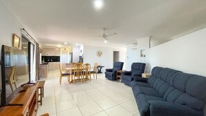A spacious living area with a large blue sofa, wooden dining table, and chairs. There is a kitchen in the background with cabinets and a refrigerator. - Seahaven - Located in Toogoom (TOOGOOM)