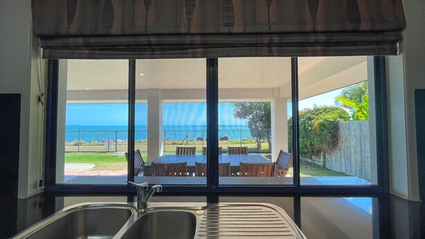 A kitchen with a large window overlooking a patio with a table and chairs, and a view of the ocean. - Seahaven - Located in Toogoom (TOOGOOM)