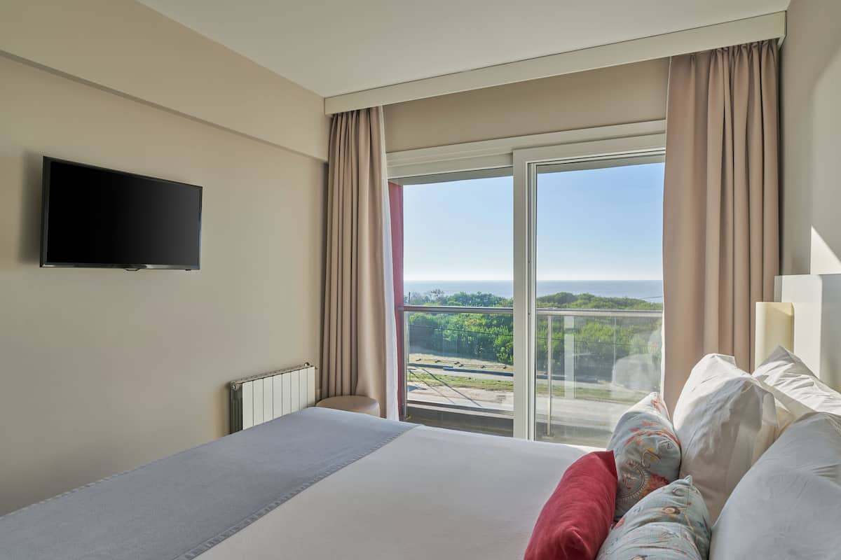 Hotelkamer met een bed, een flatscreen televisie, een groot raam met gordijnen en uitzicht op groen en een wateroppervlak.