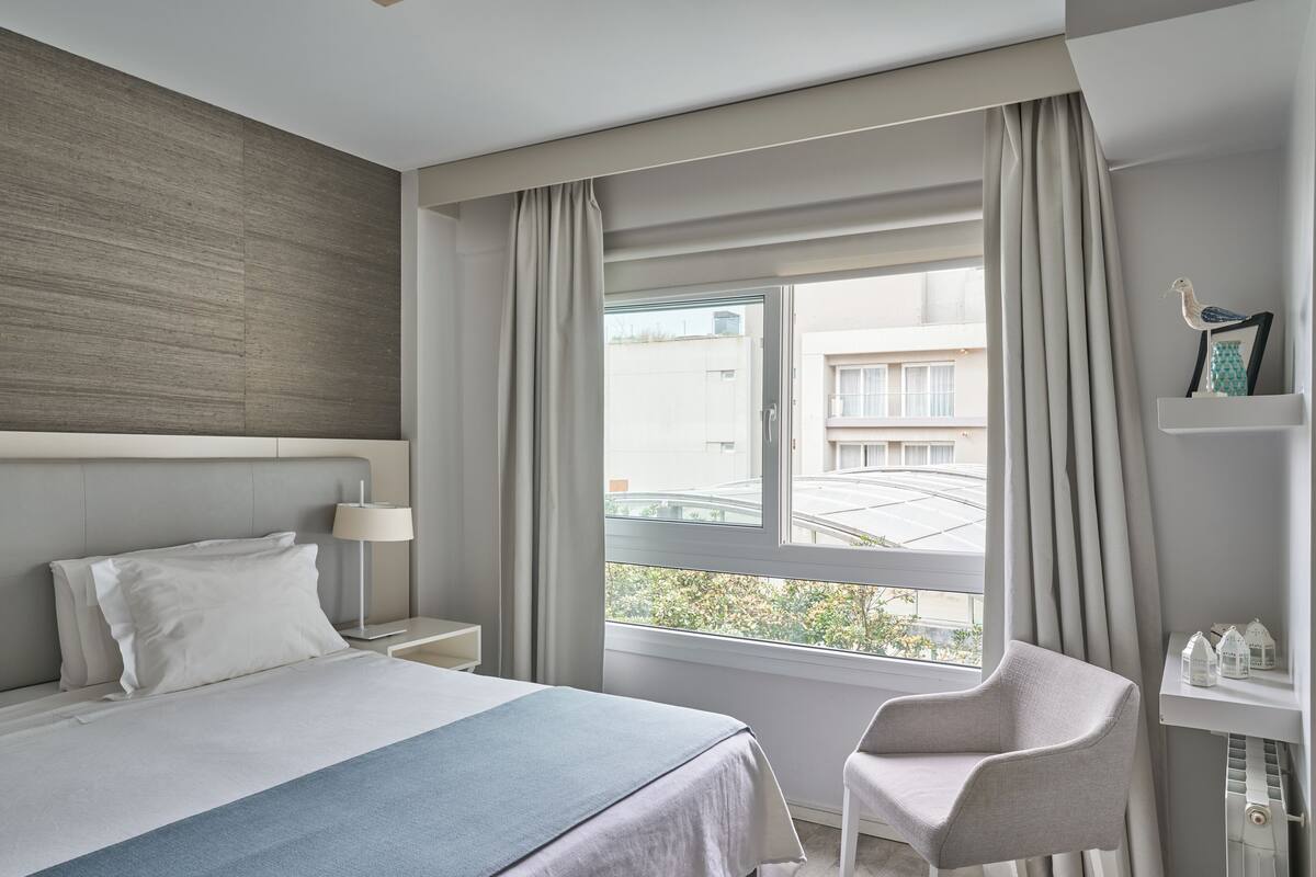 Een moderne hotelkamer met een groot bed, een fauteuil, een nachtkastje, een lamp, een raam met gordijnen en uitzicht op het omliggende landschap.
