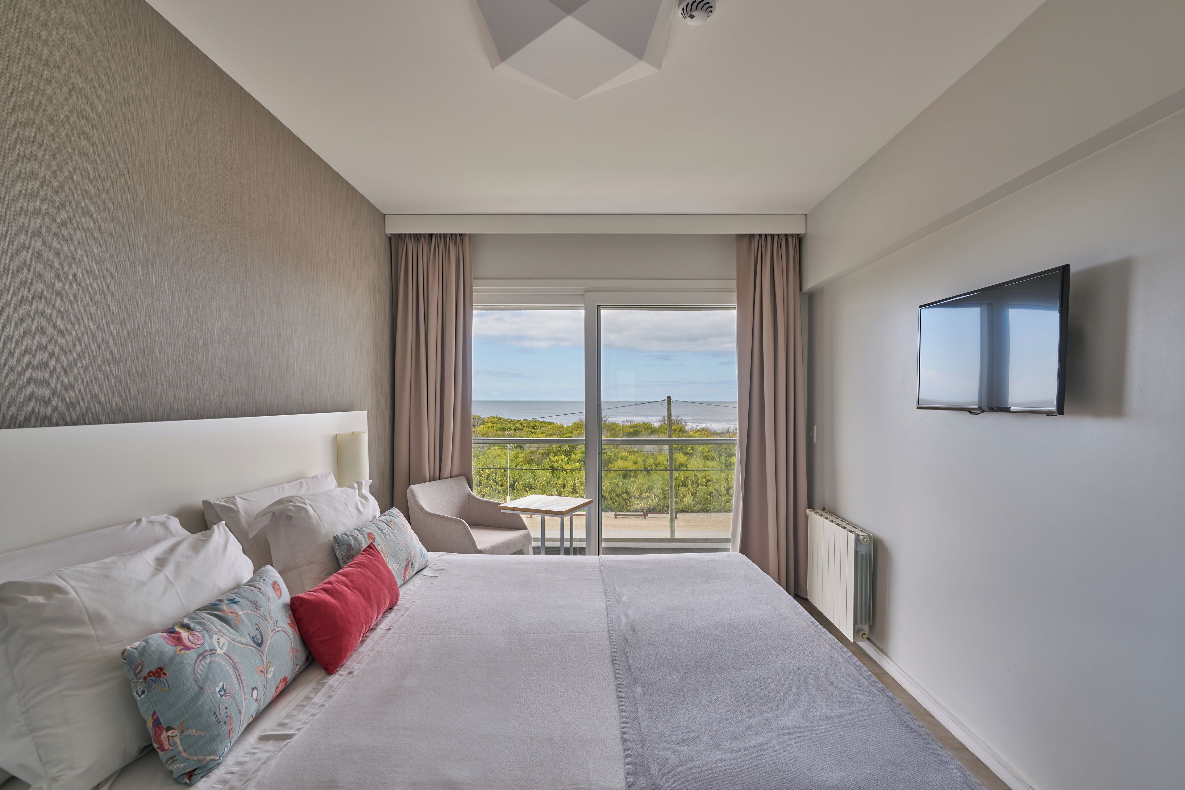 Quarto de hotel com uma cama grande, televisão na parede, varanda com vista para o verde e uma pequena mesa com cadeira.