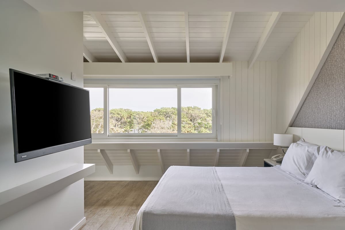 Een slaapkamer met een groot bed, een aan de muur bevestigde televisie, een raam met uitzicht op bomen en een plafond met houten balken.
