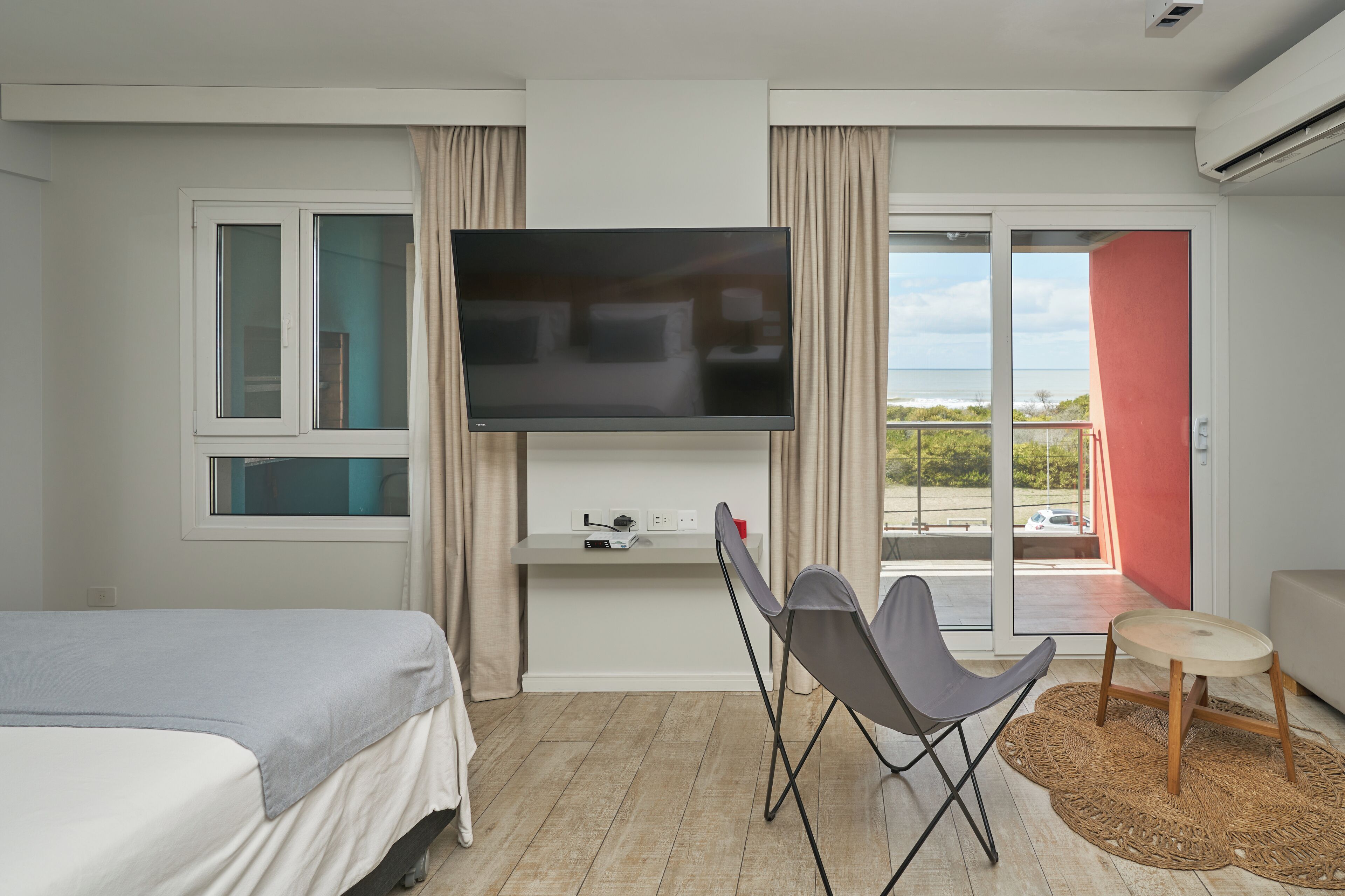 A modern hotel room with a bed, a TV, a desk with a chair, and a view through the glass door.