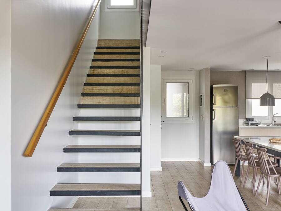 A modern kitchen with a central island, a dining area with chairs, and a staircase with a wooden handrail.