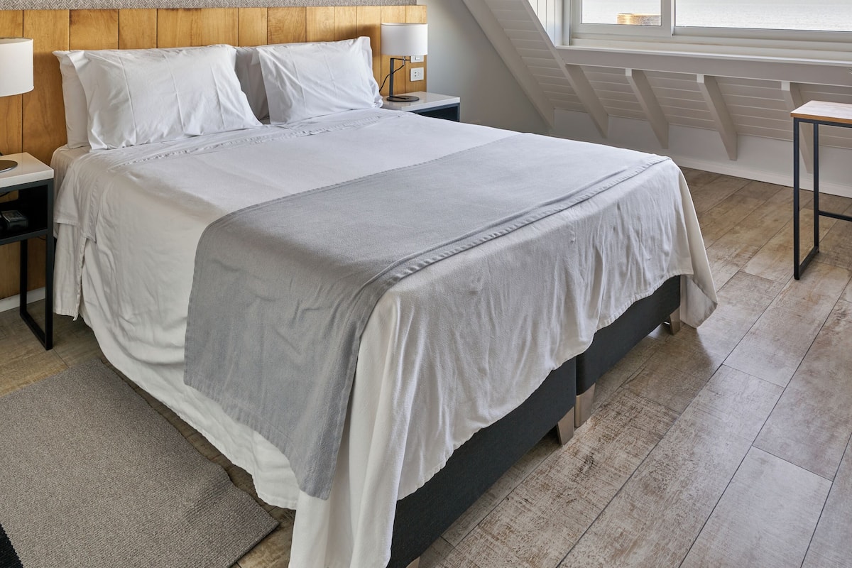 Een moderne slaapkamer met een groot bed, een houten hoofdbord, een nachtkastje met een lamp, een bureau met een stoel, en uitzicht op de zee door een groot raam.