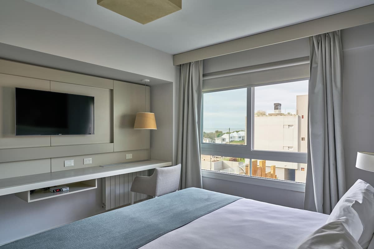 Een hotelkamer met een bed, een aan de muur bevestigde televisie, een bureau met een lamp en een raam met gordijnen.