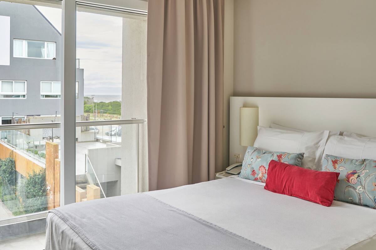 Een moderne hotelkamer met een groot bed, nachtkastje, lamp en uitzicht op een gebouw door een groot raam.