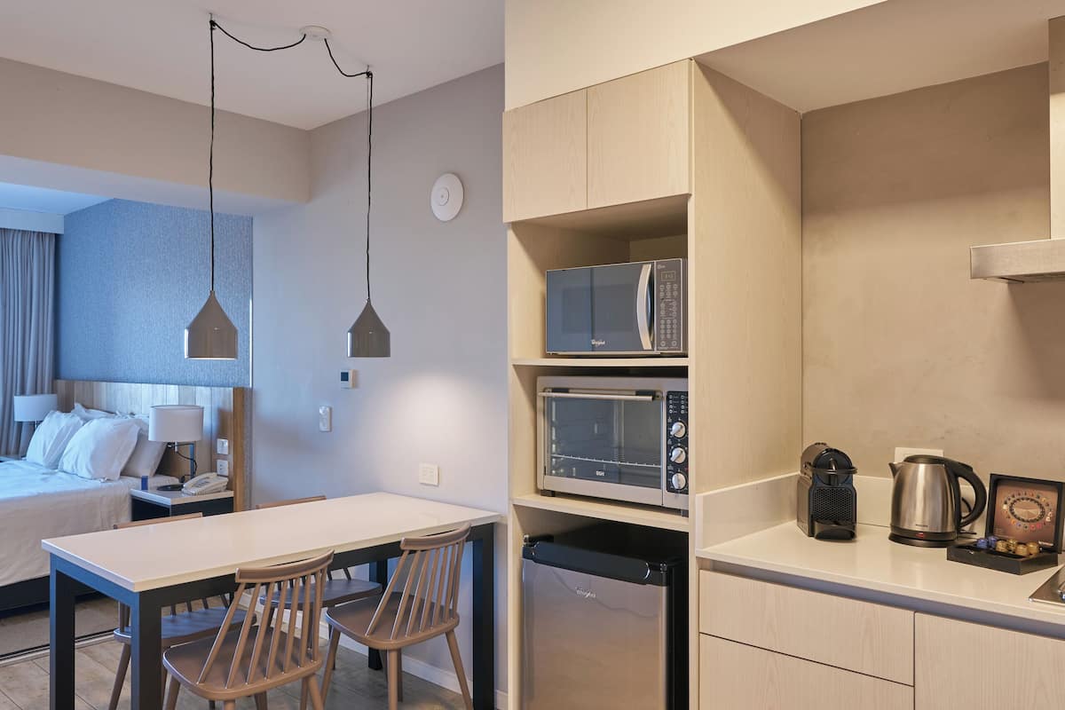 Een compacte hotelkamer met kitchenette, een zithoek met twee stoelen, een bed met nachtkastje en een groot raam.