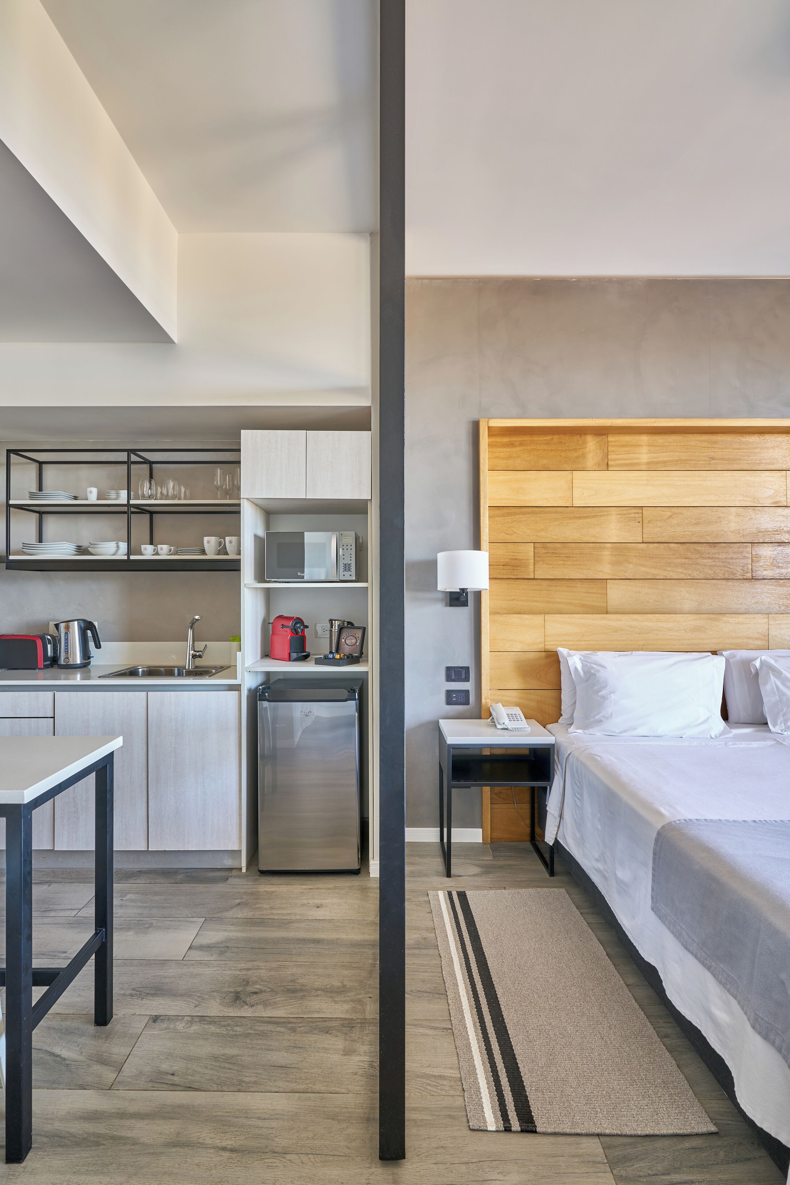 Quarto de hotel moderno com cozinha compacta, cama com roupa de cama branca, cabeceira de madeira e mesa de cabeceira com abajur.