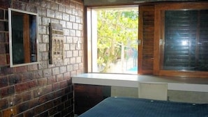A room with a brick wall, a window with a view of greenery, and a bed with a blue cover.