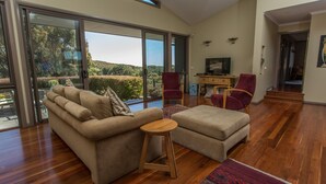 Una sala moderna con sofá, reposapiés y sillones, grandes puertas correderas de vidrio con vistas a un jardín, un escritorio de madera con televisor y una escalera que sube a otro nivel.