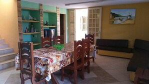 A dining area with a table covered in a patterned tablecloth, surrounded by wooden chairs. There is a television on a stand to the left, and a painting on the wall. - L'AMARELA MANSION 11 SUITES 50 BEDS NEIGHBORHOOD TO BEACH PARK (AQUIRAZ)