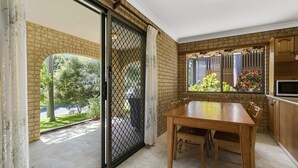 Un comedor con pared de ladrillo, puerta corredera de vidrio que da a un jardín, y una cocina con armarios de madera y microondas.