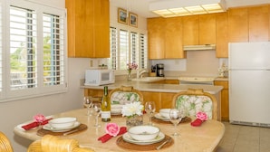Un coin repas avec une table ronde dressée pour quatre personnes, entourée de chaises en osier. On aperçoit, à l’arrière-plan, un réfrigérateur blanc et des meubles en bois.