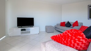 A modern living room with a flat-screen TV on a white entertainment unit, a grey sofa with red and black cushions, and a grey ottoman. - Explorer Cove Unit 6, 31 Kennedy Pde (GOLDEN BEACH)