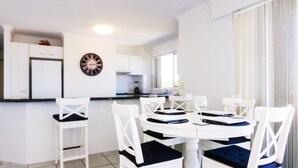 A modern kitchen with white cabinets and a clock on the wall. There is a dining area with a round table and white chairs. - Explorer Cove Unit 6, 31 Kennedy Pde (GOLDEN BEACH)