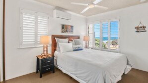 Un dormitorio con una cama, dos mesitas de noche, un ventilador de techo y vistas al océano a través de amplios ventanales.