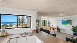 Una cocina moderna con un amplio ventanal con vista a una playa, un comedor con mesa y sillas, y una sala de estar con sofá y televisor mostrando una escena de playa.