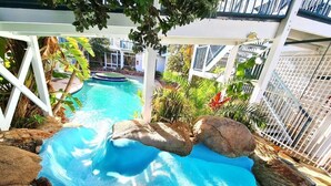 Una piscina con tobogán, rodeada de rocas y plantas, con una cerca blanca y edificios al fondo.