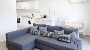 Una sala de estar moderna con un sofá seccional gris, una mesa de comedor y una zona de cocina con armarios y electrodomésticos blancos.