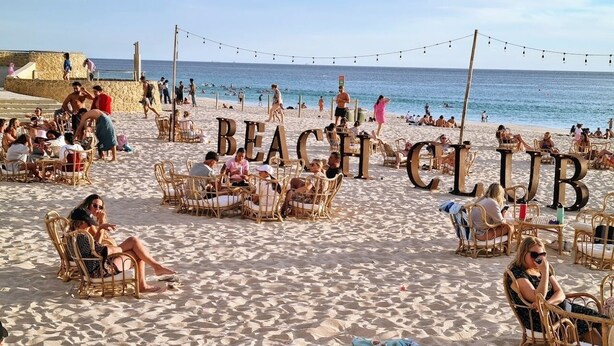 Un club de playa con sillas y mesas de mimbre, un muro de piedra y el océano al fondo.