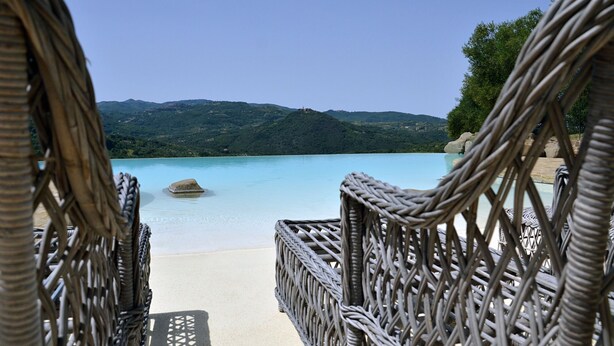 Des chaises en osier sont disposées face à une piscine à l’eau cristalline, et en arrière-plan, on aperçoit des collines verdoyantes.