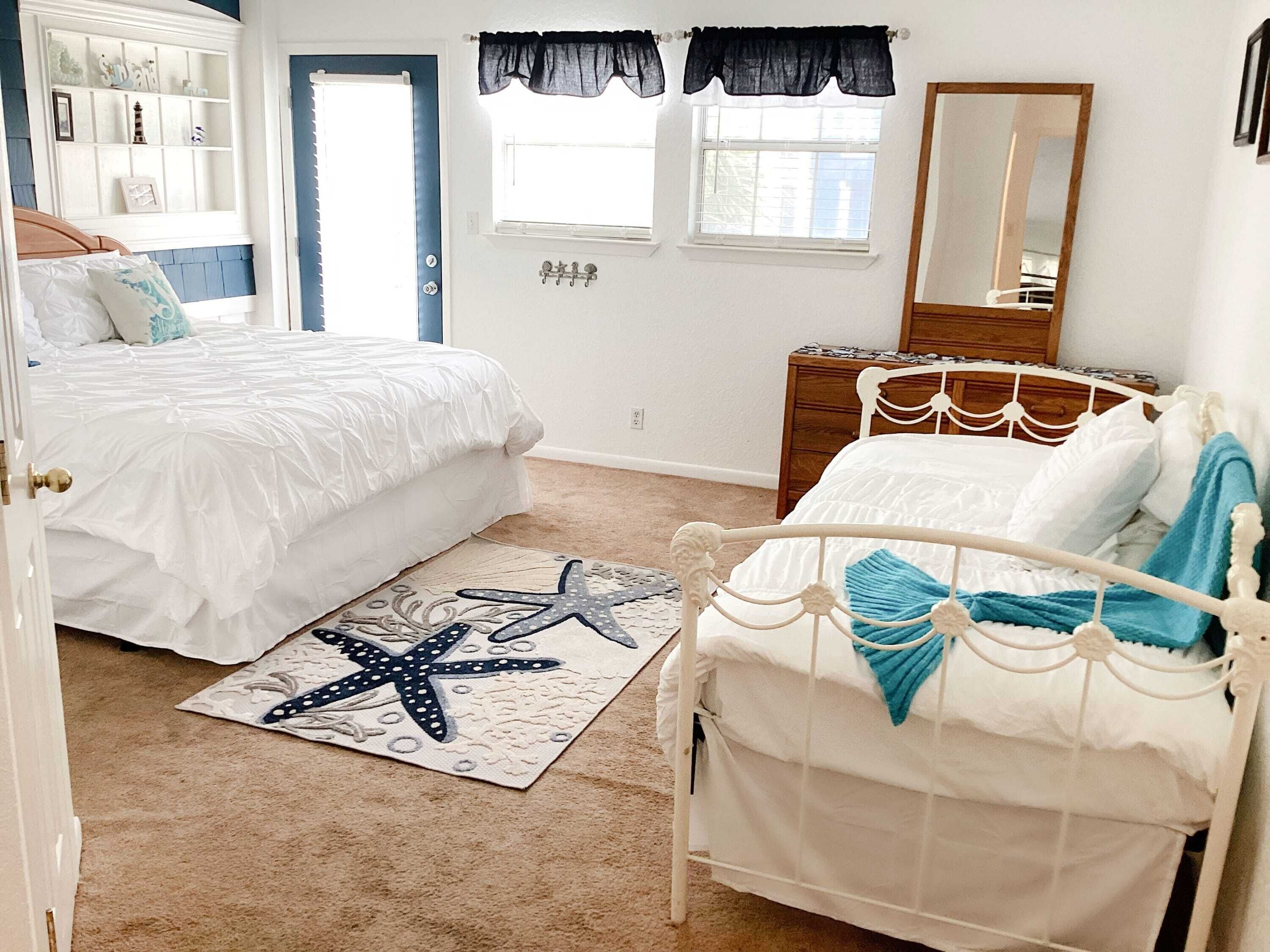 A bedroom with two beds, a starfish rug, a dresser, a mirror, and a window with black curtains.