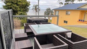 A wicker outdoor seating area with a glass-top table, overlooking a body of water and a yellow building. - SeaEsta - Waterfront - Jervis Bay  - 5 minutes to Hyams Beach (Erowal Bay)