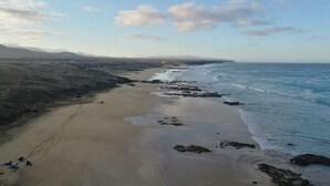 Een kustlandschap met een zandstrand, een rotsachtige kustlijn en verafgelegen bergen onder een gedeeltelijk bewolkte lucht.