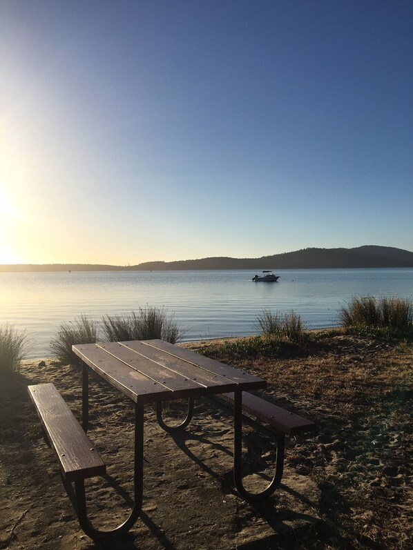 Una mesa de picnic de madera con bancos en una playa arenosa, con vistas a un lago tranquilo con un barco a lo lejos y colinas al fondo.