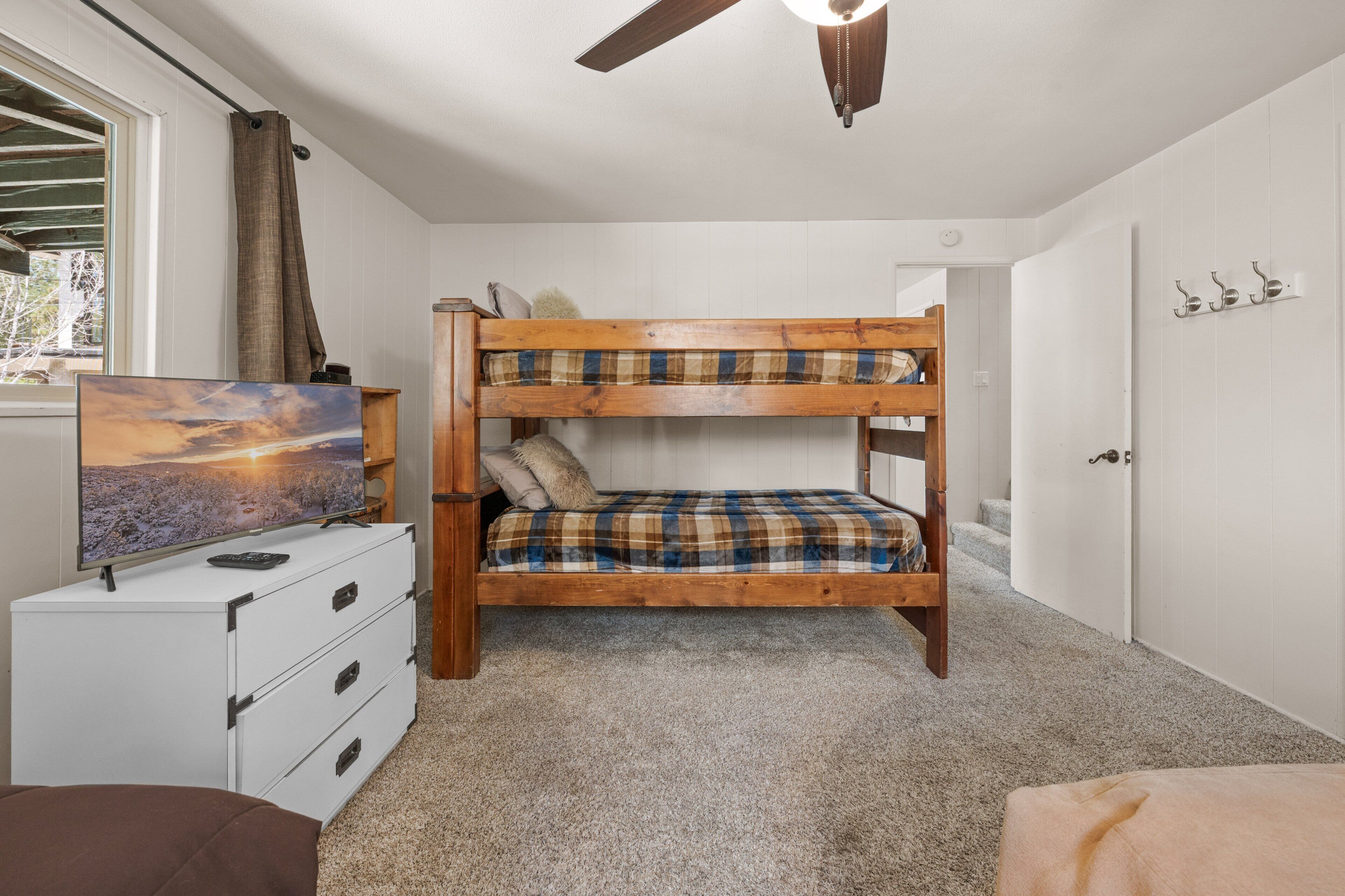 A bunk bed with a plaid mattress, a television on a white dresser, and a ceiling fan.