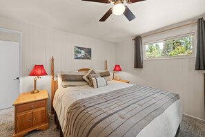 Une chambre à coucher avec un lit, deux tables de chevet avec des lampes, un ventilateur de plafond, une fenêtre avec des rideaux et un tableau accroché au mur.